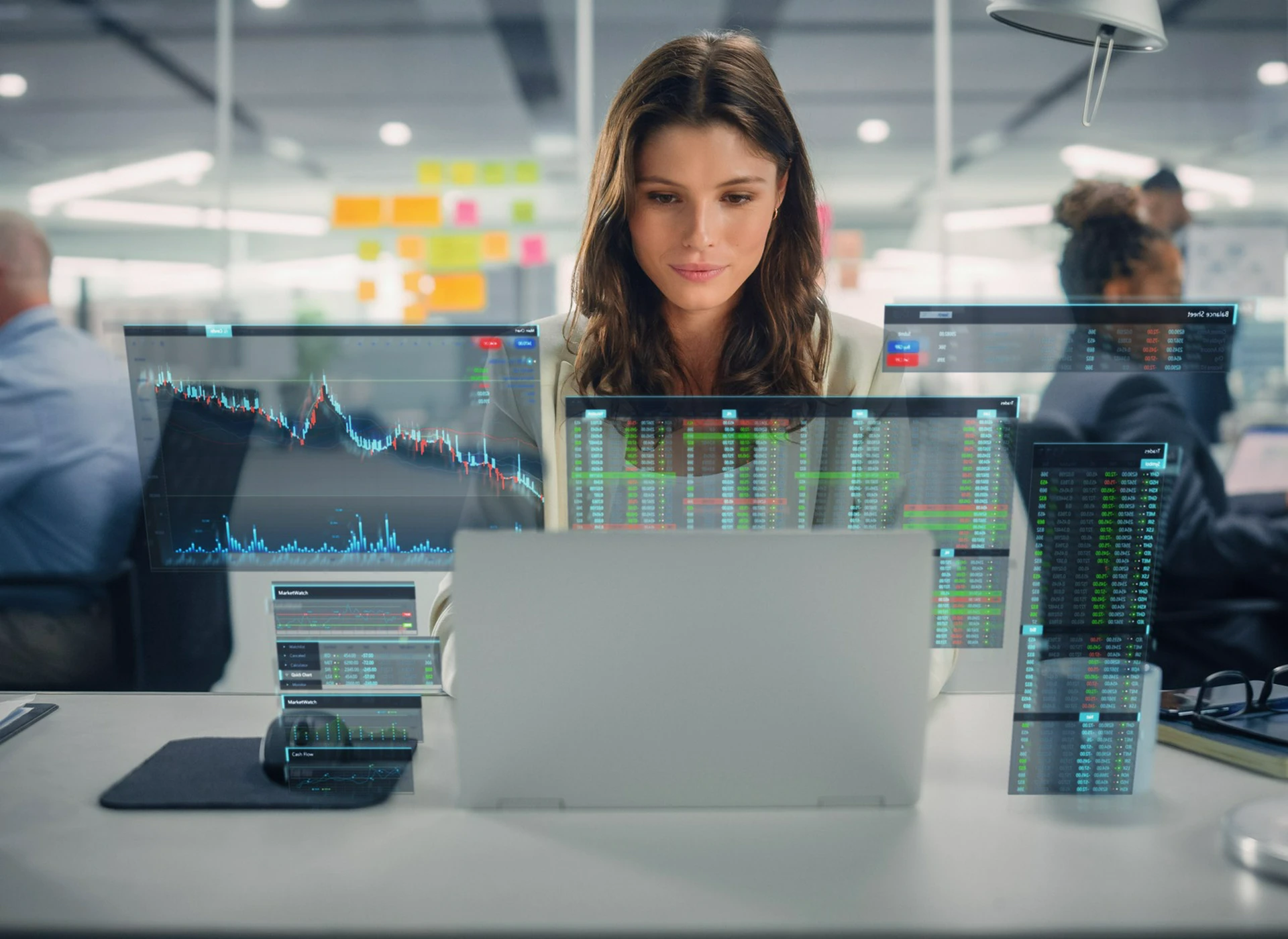 Woman working on a&nbsp;laptop surrounded by&nbsp;transparent digital screens displaying financial charts and market data